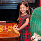 A young girl in a red Paces Mill Plaid dress kneels on a green chair, smiling beside a wooden checkers board, her look finished with a T.B.B.C. x Lele Sadoughi Knotted Headband by The Beaufort Bonnet Company.