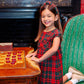 A young girl in The Beaufort Bonnet Company's Girls' Darcy Dress kneels on a green striped chair, smiling beside a wooden checkers board in a richly decorated room.