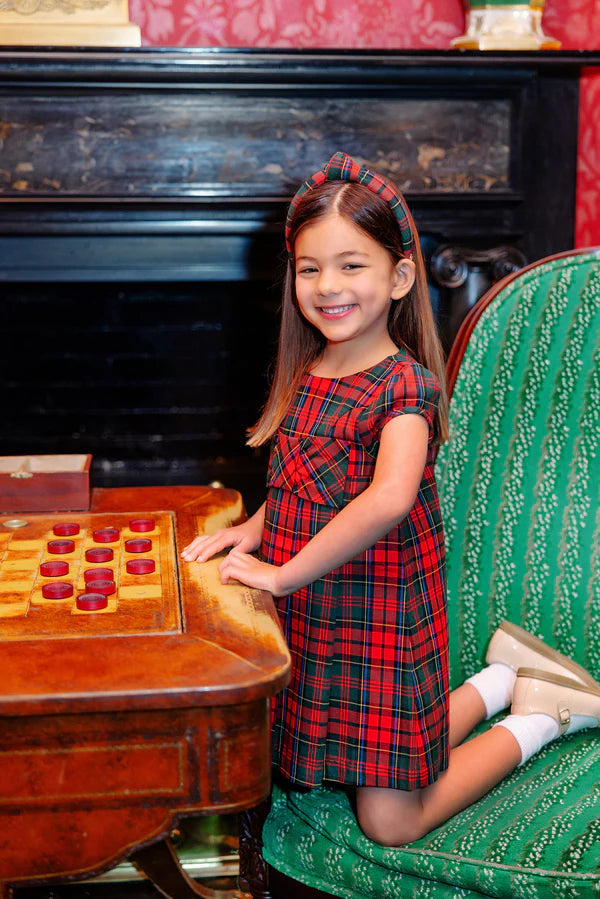 A young girl in The Beaufort Bonnet Company's Girls' Darcy Dress kneels on a green striped chair, smiling beside a wooden checkers board in a richly decorated room.