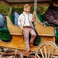 A young boy wearing The Beaufort Bonnet Company's Baby Boys' and Boys' Sherpa Van Camp Vest stands on a vintage horse-drawn carriage step, smiling at the camera, while an adult driver with a hat sits at the front.
