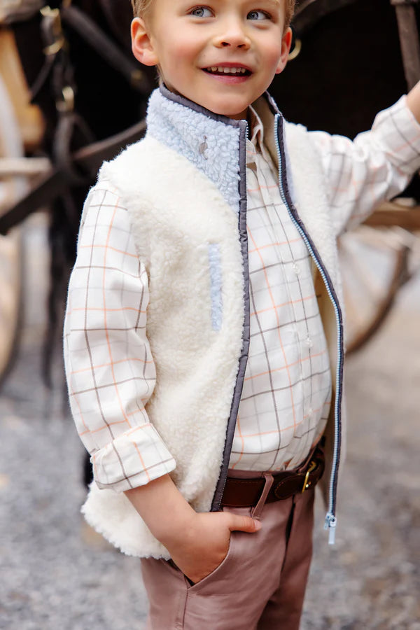 A young boy in The Beaufort Bonnet Company's Baby Boys' and Boys' Sherpa Van Camp Vest, layered over a plaid shirt and pink pants, stands outdoors with one hand in his pocket, smiling and looking to the side.