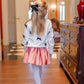 A young girl with long blonde hair, dressed in a white sweater with black bows, pink skirt, white tights, and black shoes, wears the T.B.B.C. x Lele Sadoughi Britney Barrette by The Beaufort Bonnet Company as she stands indoors, facing away.