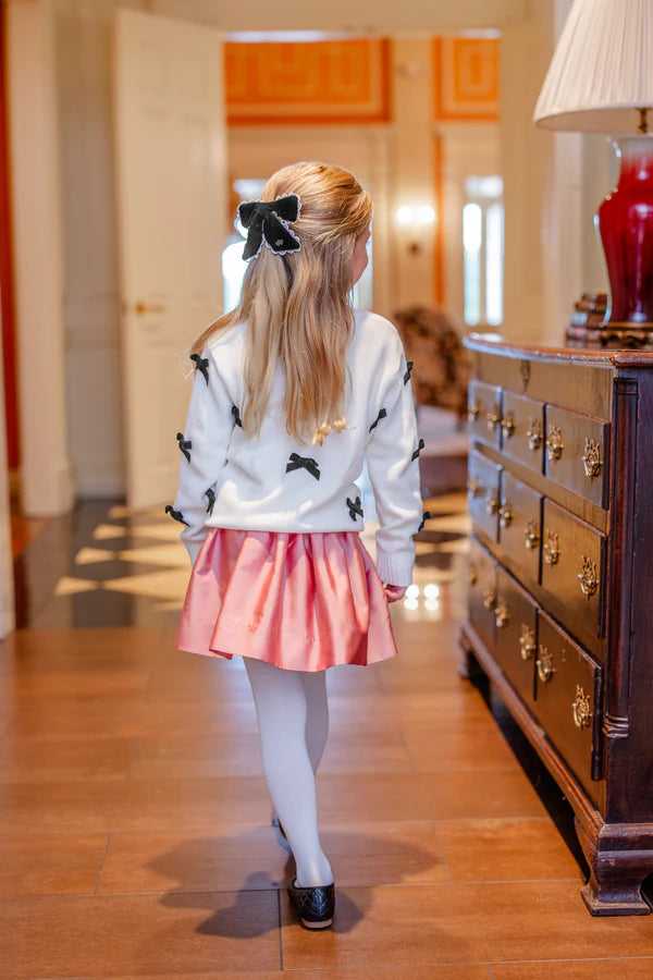 A young girl with long blonde hair, dressed in a white sweater with black bows, pink skirt, white tights, and black shoes, wears the T.B.B.C. x Lele Sadoughi Britney Barrette by The Beaufort Bonnet Company as she stands indoors, facing away.