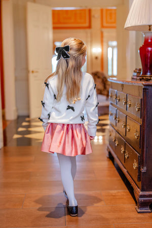 A young girl with long blonde hair, dressed in a white sweater with black bows, pink skirt, white tights, and black shoes, wears the T.B.B.C. x Lele Sadoughi Britney Barrette by The Beaufort Bonnet Company as she stands indoors, facing away.