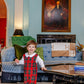 A smiling young child wears the Baby Boys' Lawson Longall by The Beaufort Bonnet Company, standing in a stylish living room with a blue sofa and a framed portrait on the wall.