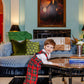 A young boy in The Beaufort Bonnet Company’s Baby Boys' Lawson Longall leans on a coffee table in a stylish living room with blue walls, a patterned sofa, and a portrait above the fireplace.