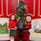 A young girl in The Beaufort Bonnet Company Girls' Critter Beasley Bag Skirt decorates a small Christmas tree on a table between two armchairs, with ornaments and decorations nearby.