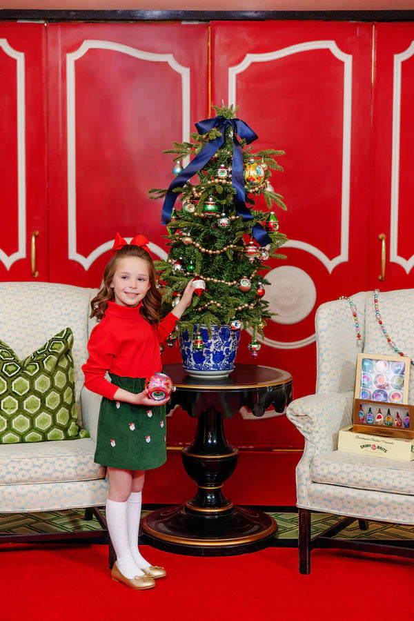 A young girl in The Beaufort Bonnet Company Girls' Critter Beasley Bag Skirt decorates a small Christmas tree on a table between two armchairs, with ornaments and decorations nearby.