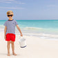 A toddler boy in sunglasses, a striped shirt, and The Beaufort Bonnet Company Baby Boys' Sheffield Shorts stands barefoot on a sandy beach near the ocean, holding a white bucket.