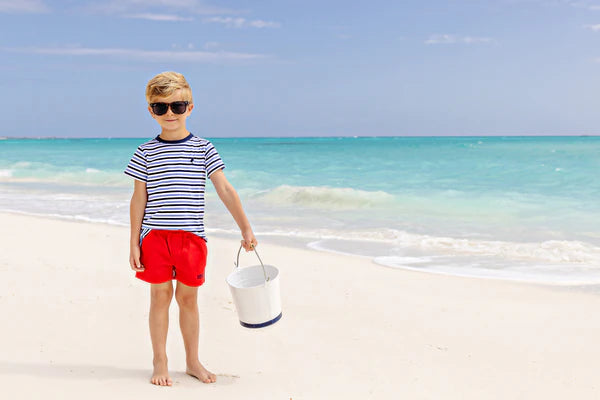 A toddler boy in sunglasses, a striped shirt, and The Beaufort Bonnet Company Baby Boys' Sheffield Shorts stands barefoot on a sandy beach near the ocean, holding a white bucket.