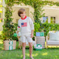 A barefoot boy in Boys' Prepletic™ Charlie's Chinos by The Beaufort Bonnet Company holds a green bocce ball on a grassy lawn, with bocce balls and a seating area in the background.