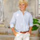 Young boy with blond hair wears The Beaufort Bonnet Company Boys' Prepletic™ Charlie's Chinos and a light blue shirt, smiling outdoors with hands in his pockets by a weathered wall and window.