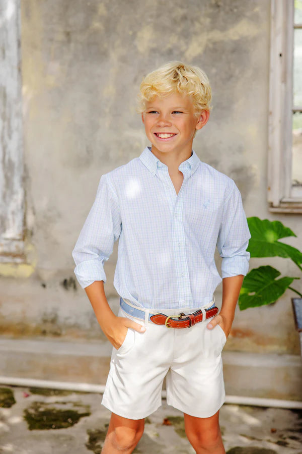 Young boy with blond hair wears The Beaufort Bonnet Company Boys' Prepletic™ Charlie's Chinos and a light blue shirt, smiling outdoors with hands in his pockets by a weathered wall and window.
