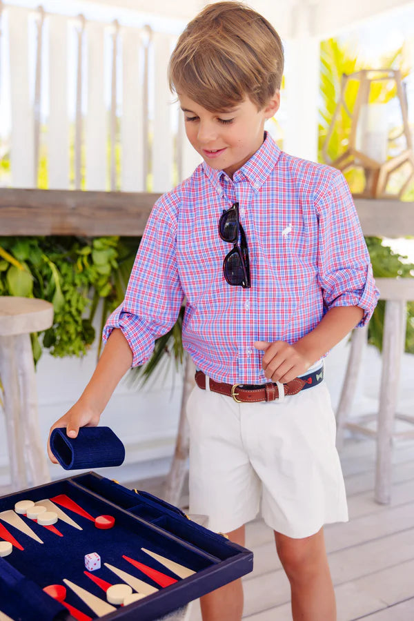 A young boy in a plaid shirt and white Boys' Prepletic™ Charlie's Chinos by The Beaufort Bonnet Company plays backgammon indoors, holding a dice cup above the game board.