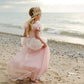 A young girl in a Great Pretenders Antique Princess Gown and crown walks barefoot on the sandy beach near the water, with waves and a cloudy sky behind her.