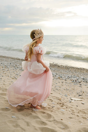A young girl in a Great Pretenders Antique Princess Gown and crown walks barefoot on the sandy beach near the water, with waves and a cloudy sky behind her.