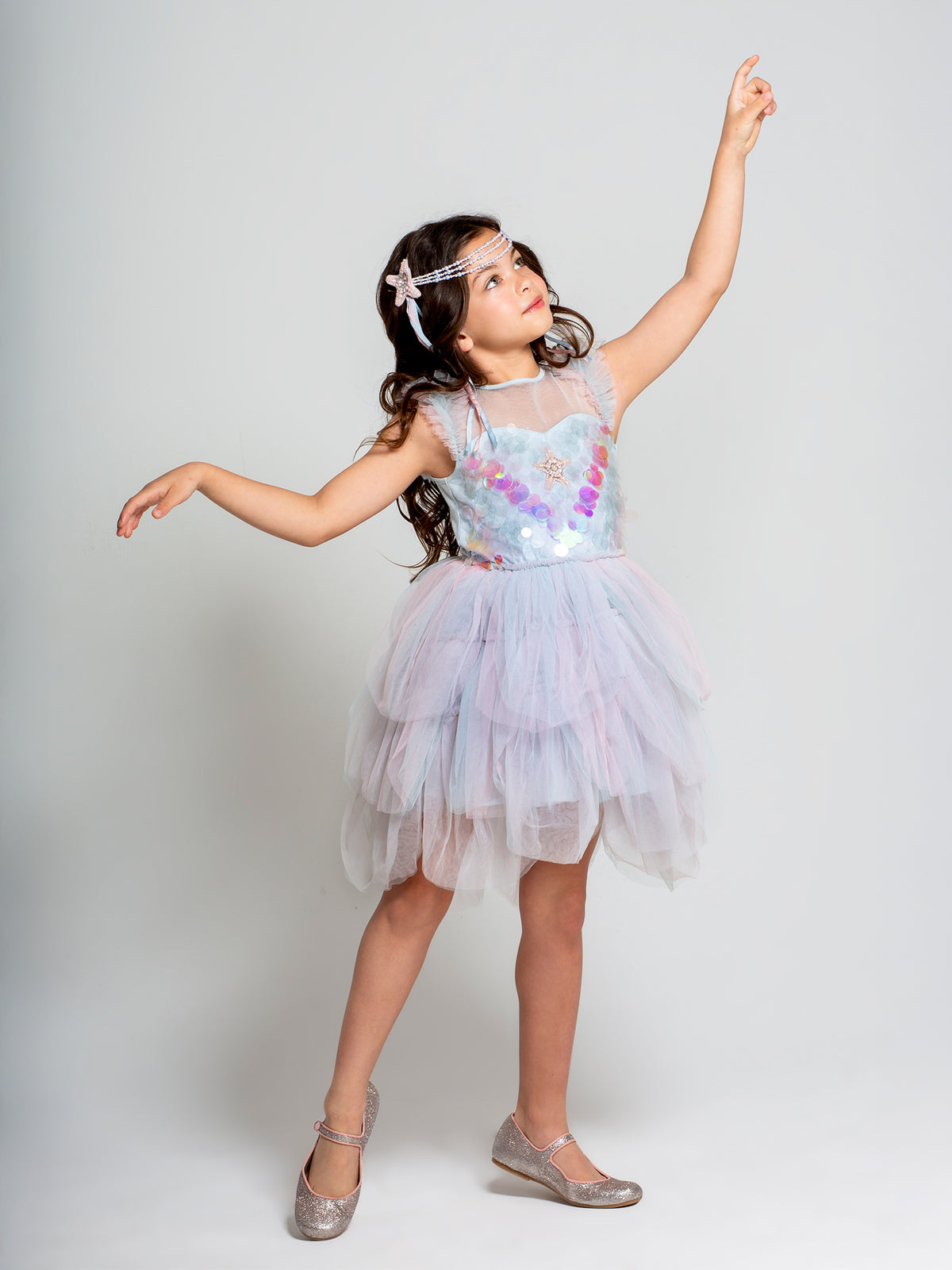 A young girl in the Tutu du Monde Girls’ Mermaid Magic Tutu Dress and matching headband poses with one arm raised, balancing on one foot against a soft, light background. A delicate sequin starfish accent brings an enchanting, ocean-inspired charm to her whimsical ensemble.
