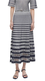Person in a Simkhai Larken Midi Skirt featuring a bold checkered pattern, pleats, and paired with black strappy sandals.