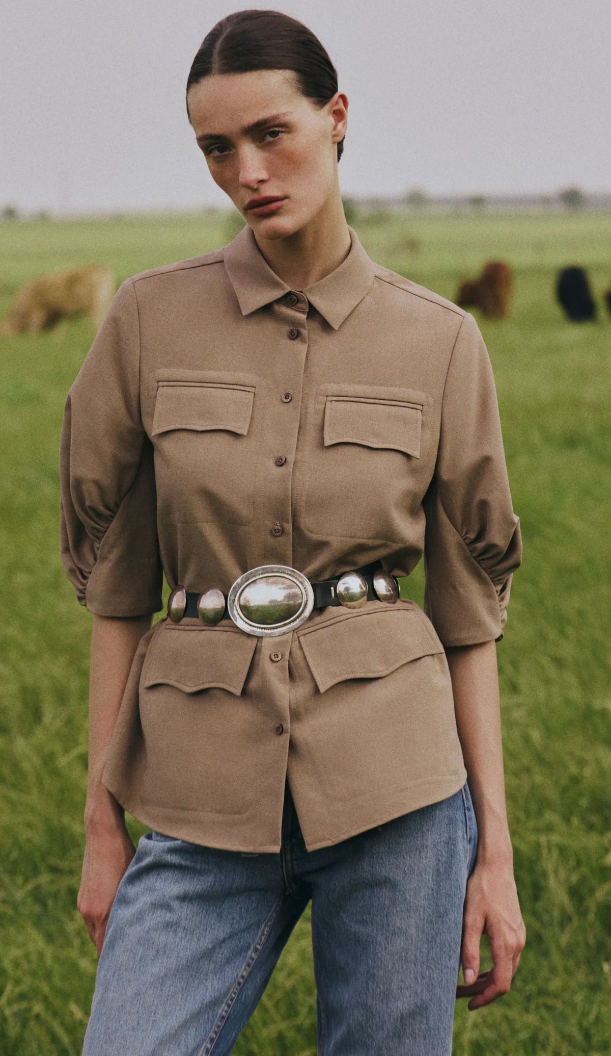 A person stands in a grassy field wearing the Lela Rose Melange Crepe Camp Shirt, which features a classic silhouette. They have paired it with jeans and a large belt adorned with silver details, while cows graze in the background.