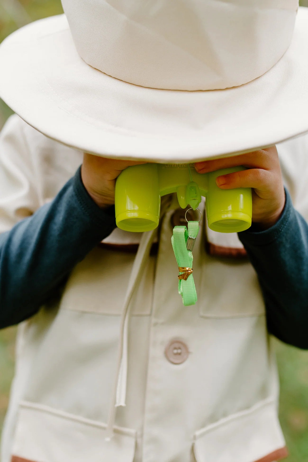 A child dressed in the Great Pretenders Forest Guardian Set with Accessories, including a cream safari costume and hat, holds green toy binoculars up to their face, covering their eyes.