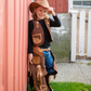 A child dressed in the Great Pretenders Cowboy Vest and Chaps, size 7-8, leans against a red barn, smiling while wearing a brown cowboy hat and jeans.