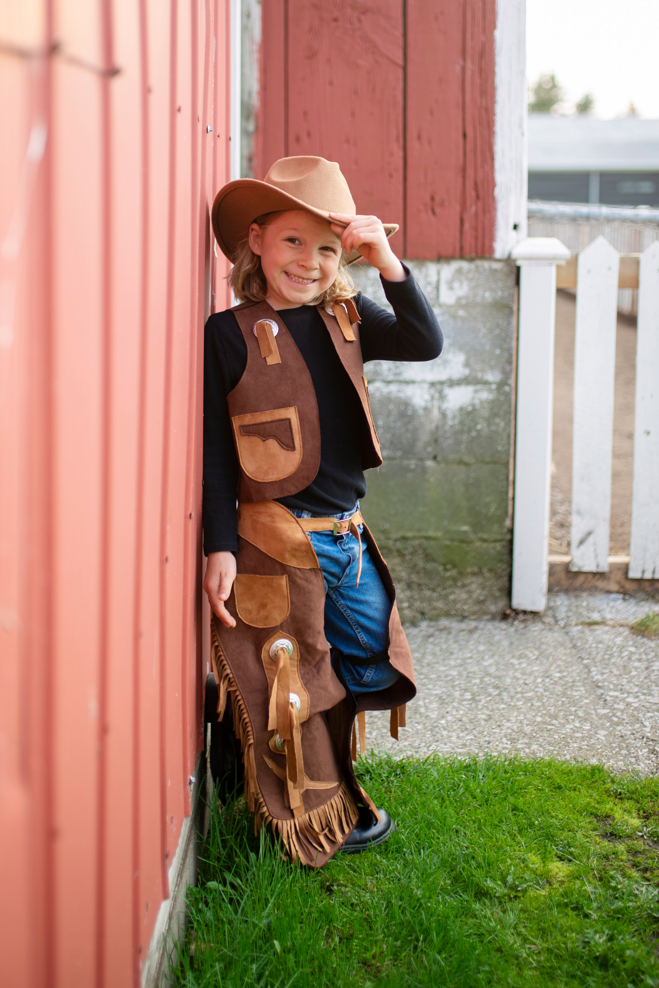 A child dressed in the Great Pretenders Cowboy Vest and Chaps, size 7-8, leans against a red barn, smiling while wearing a brown cowboy hat and jeans.