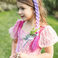 A young girl stands on the grass, holding a wand, wearing a pink princess dress with sheer sleeves and the Great Pretenders Mermazing Mermaid Hair Braid in purple.