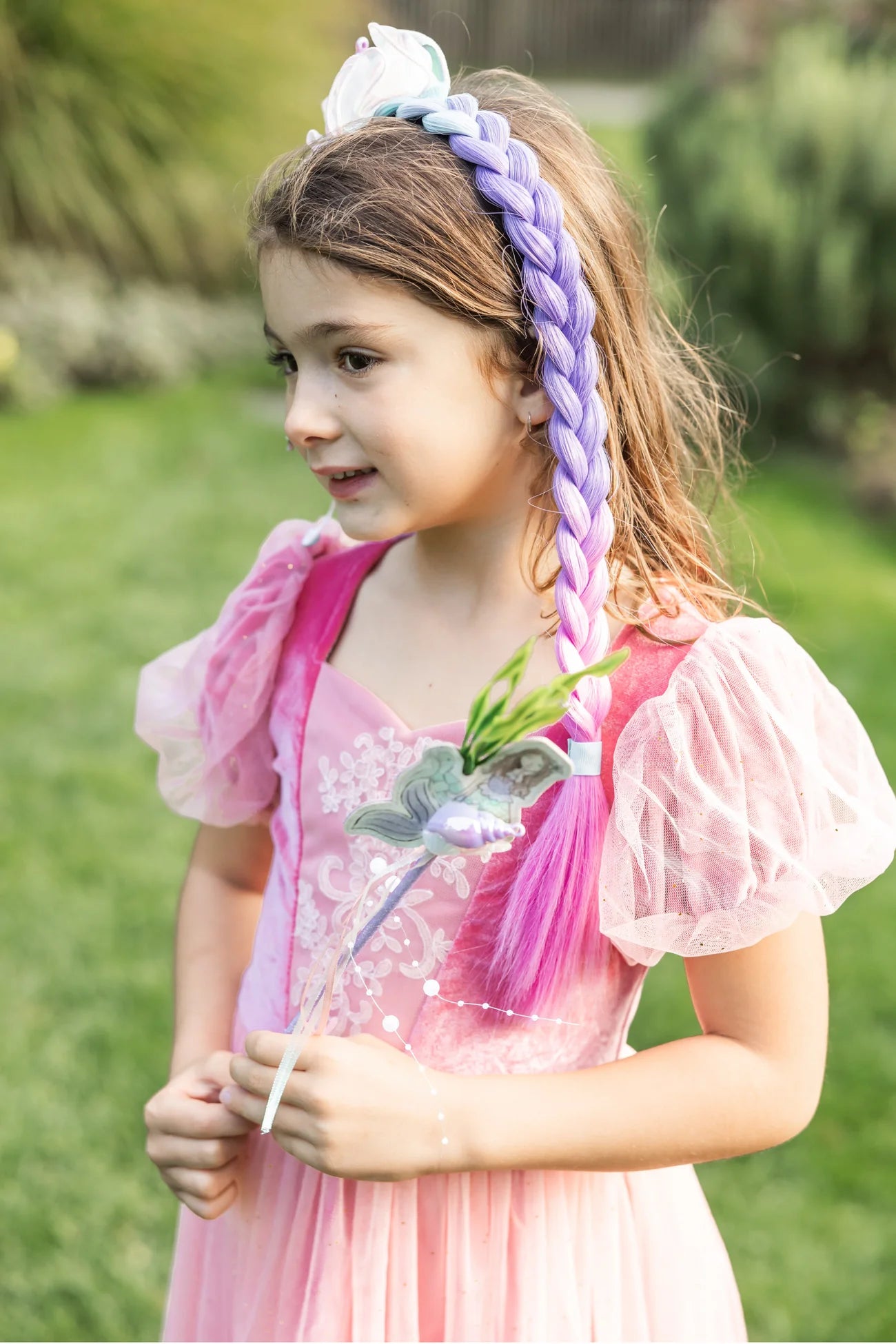 A young girl stands on the grass, holding a wand, wearing a pink princess dress with sheer sleeves and the Great Pretenders Mermazing Mermaid Hair Braid in purple.