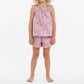 A young child is wearing the Mer Girls’ Colette Top and Shorts Set, a pink floral cotton outfit with a sleeveless top and coordinating fringe-trim shorts, standing barefoot against a plain white background.