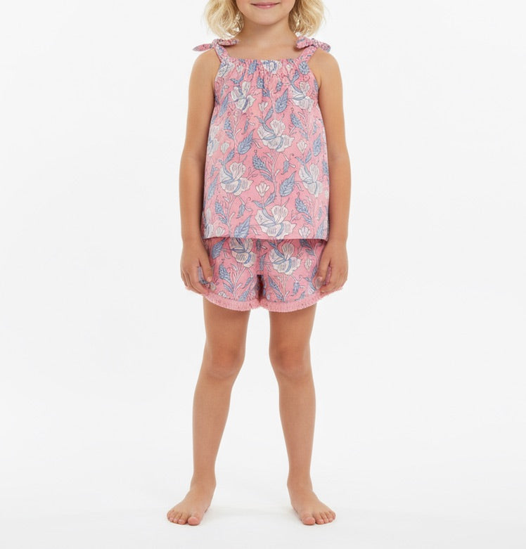 A young child is wearing the Mer Girls’ Colette Top and Shorts Set, a pink floral cotton outfit with a sleeveless top and coordinating fringe-trim shorts, standing barefoot against a plain white background.