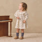 A young child wearing the Quincy Mae Baby Girls’ and Girls’ Long Sleeve Isla Dress stands on a textured rug next to a wooden piano, looking to the side.