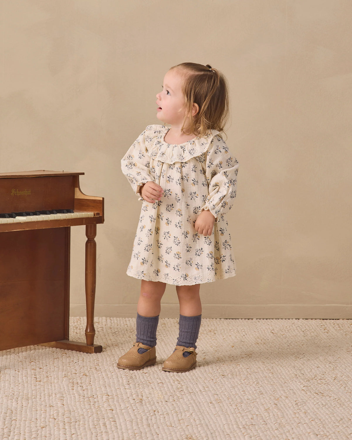 A young child wearing the Quincy Mae Baby Girls’ and Girls’ Long Sleeve Isla Dress stands on a textured rug next to a wooden piano, looking to the side.