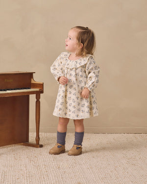 A young child wearing the Quincy Mae Baby Girls’ and Girls’ Long Sleeve Isla Dress stands on a textured rug next to a wooden piano, looking to the side.