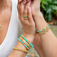 A person in a white top wears gemstone jewelry—Juliska Capucine De Wulf’s Berry Gem Hinged Bangle, a turquoise necklace, and an elegant gold ring—set against lush greenery.