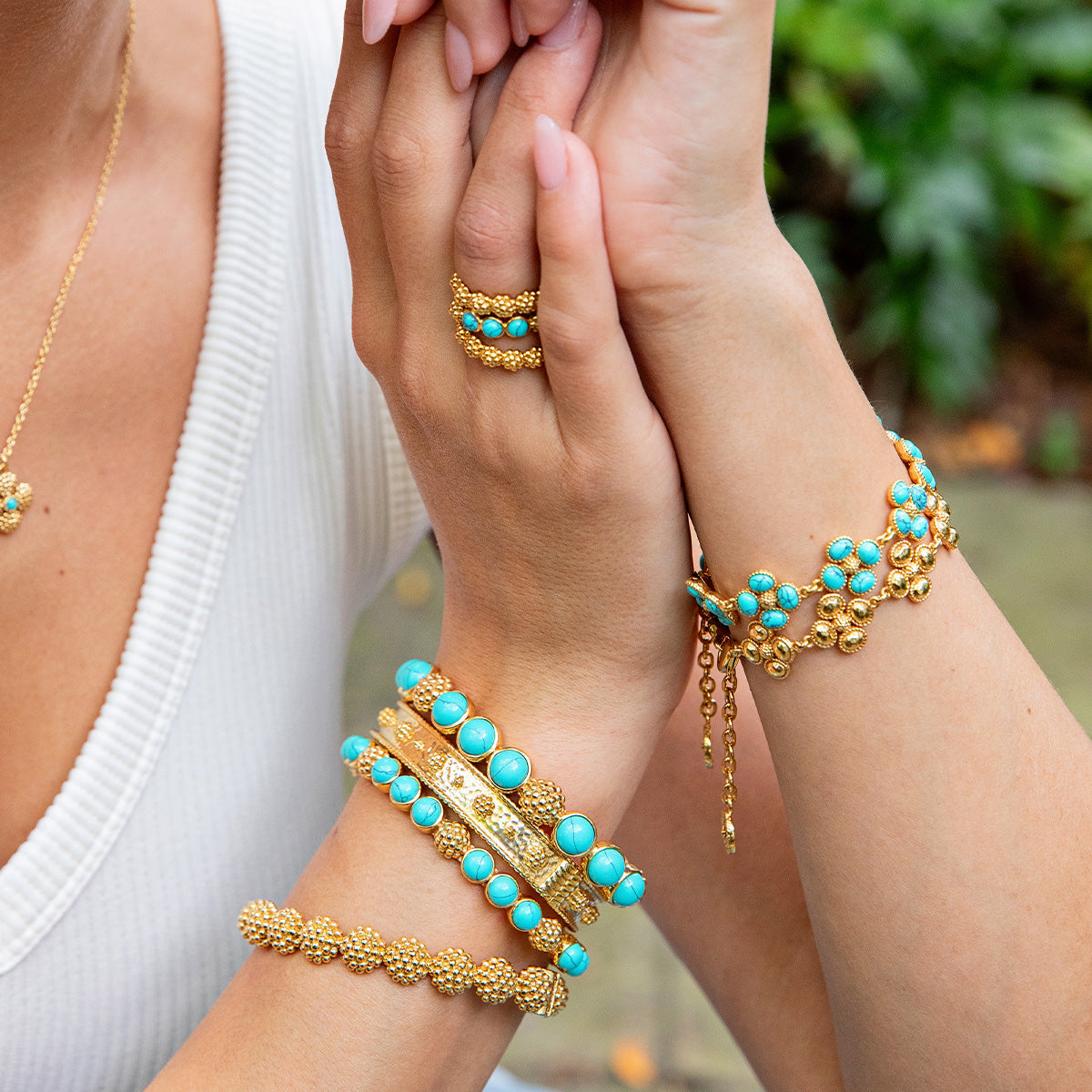 A person in a white top wears gemstone jewelry—Juliska Capucine De Wulf’s Berry Gem Hinged Bangle, a turquoise necklace, and an elegant gold ring—set against lush greenery.