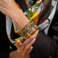 A person in dark clothing and a patterned silk scarf showcases Juliska Capucine De Wulf's Berry Gem Hinged Bangle on their wrist—its gold finish and green gemstones evoke the elegance of regal jewelry.