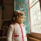A young child stands by a window in the Louise Misha Girls’ Josia Cardigan, a cream and pink cotton knit featuring button closure, with soft daylight and a painted floral panel in the background.