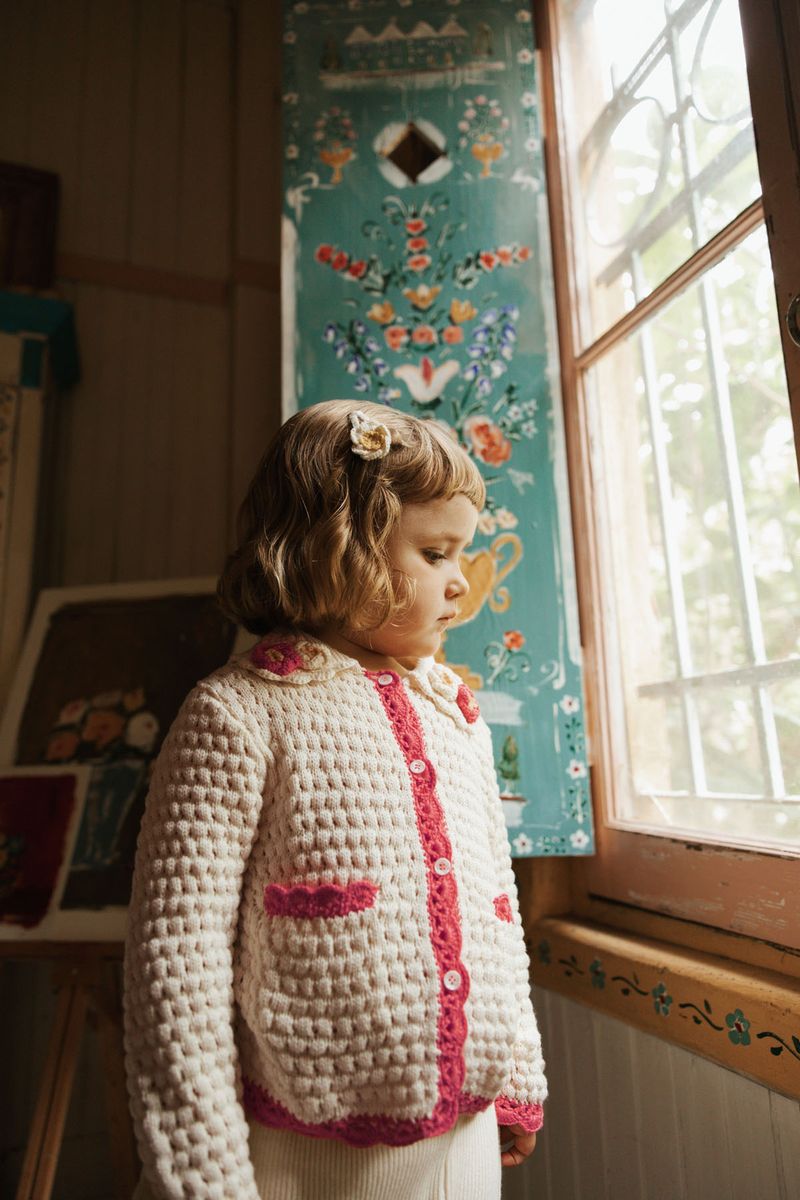 A young child stands by a window in the Louise Misha Girls’ Josia Cardigan, a cream and pink cotton knit featuring button closure, with soft daylight and a painted floral panel in the background.