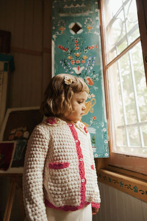 A young child stands by a window in the Louise Misha Girls’ Josia Cardigan, a cream and pink cotton knit featuring button closure, with soft daylight and a painted floral panel in the background.