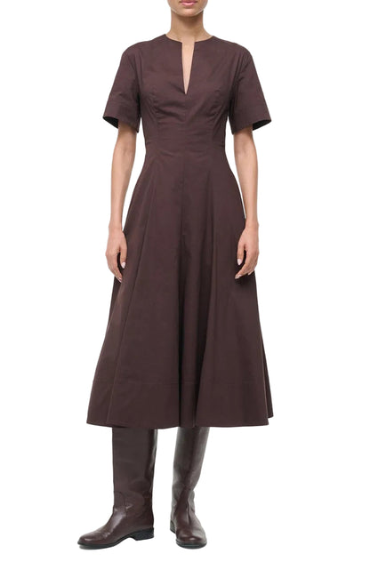 A person wearing the Staud Frances Dress, a brown split neck poplin dress with an a-line skirt, and brown knee-high boots, stands against a white background.