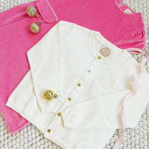 A Baby Girls' and Girls' Gold Button Cambridge Cardigan by The Beaufort Bonnet Company and a pink velvet dress are displayed on a textured surface, accented with gold ornaments and a pink ribbon.