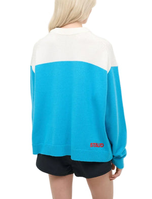 A person with long blonde hair, facing away from the camera, wears a blue and white Staud Reya Sweater featuring red "STAUD" text on the back, styled with black shorts.