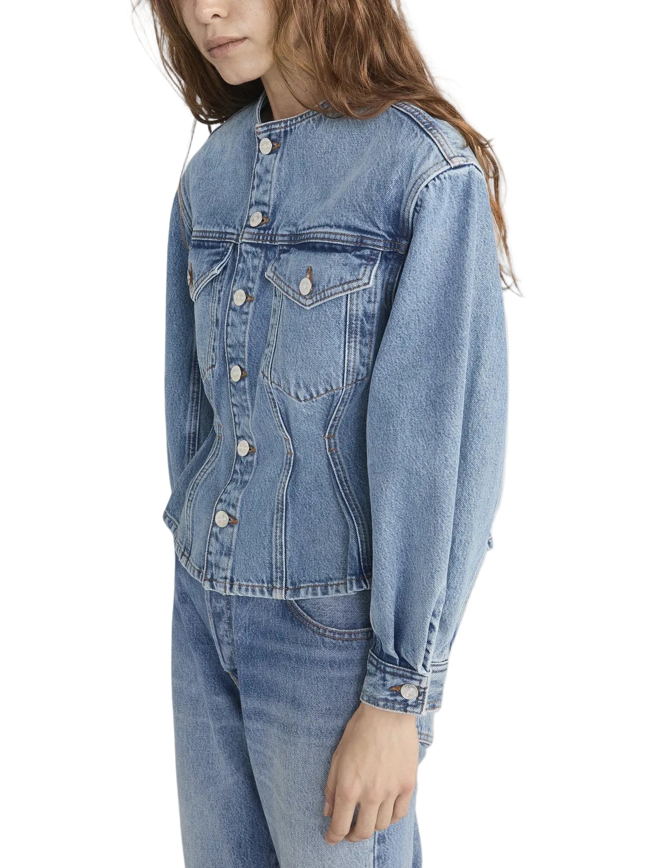 A person with long brown hair models Frame's The Nipped Collarless Jacket, paired with matching denim jeans, standing against a white background.