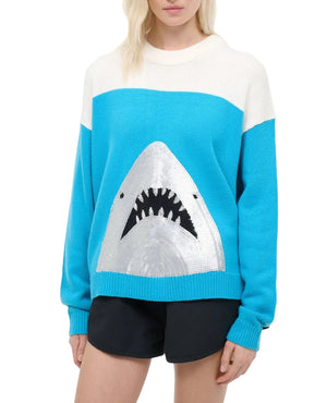 A person wears the Staud Reya Sweater, a blue and white lightweight knit featuring a large shark face on the front, styled with black shorts.