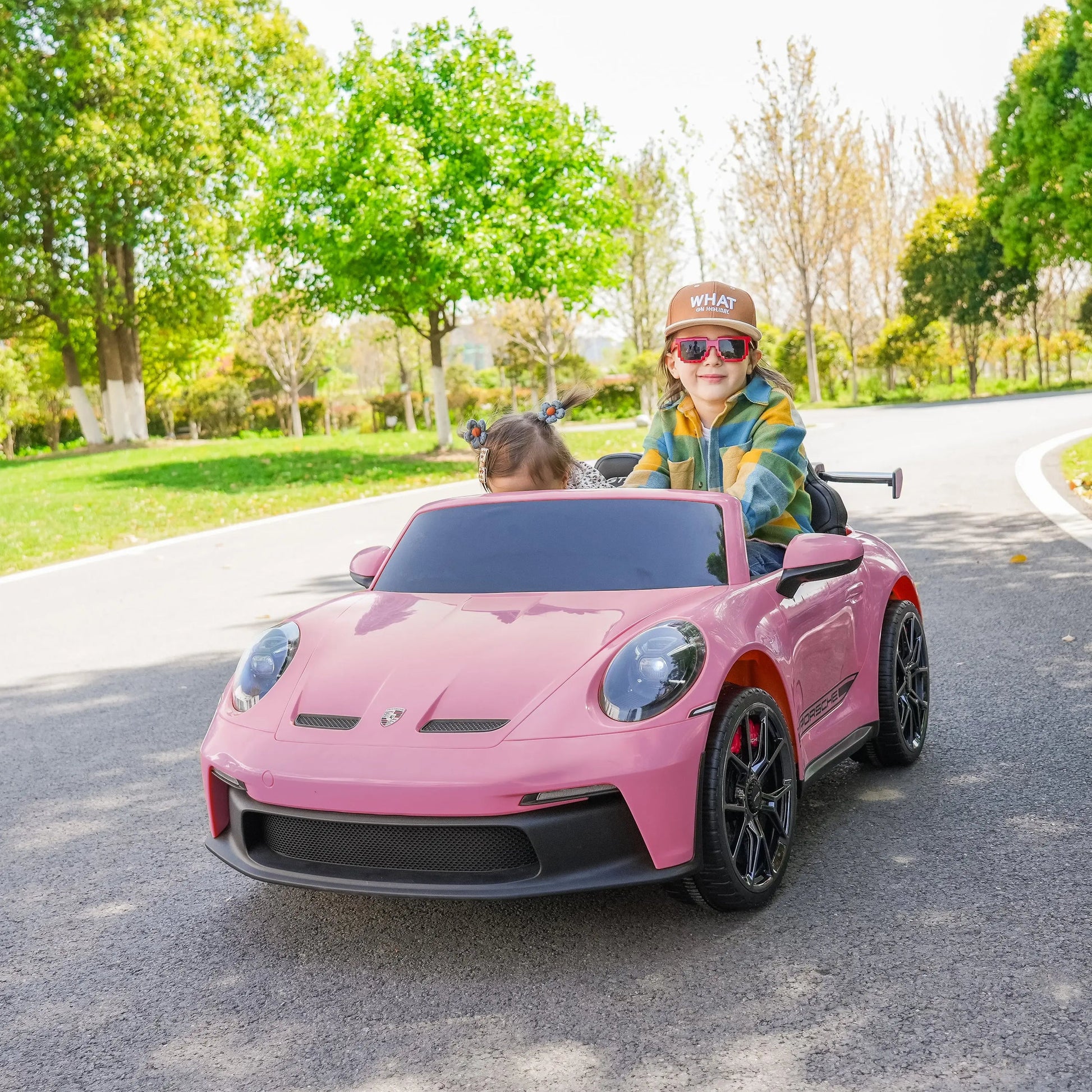 Two children ride in a Freddo 24V Porsche 911 GT3 Pink 2 Seater Ride-On on a paved path in a park, surrounded by green trees and grass under a clear sky.