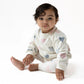 A baby sits on a white surface wearing the Angel Dear French Terry Puffy Long Sleeved Oversized Sweatshirt + Legging set with pink, blue, and gray heart patterns, looking toward the camera.