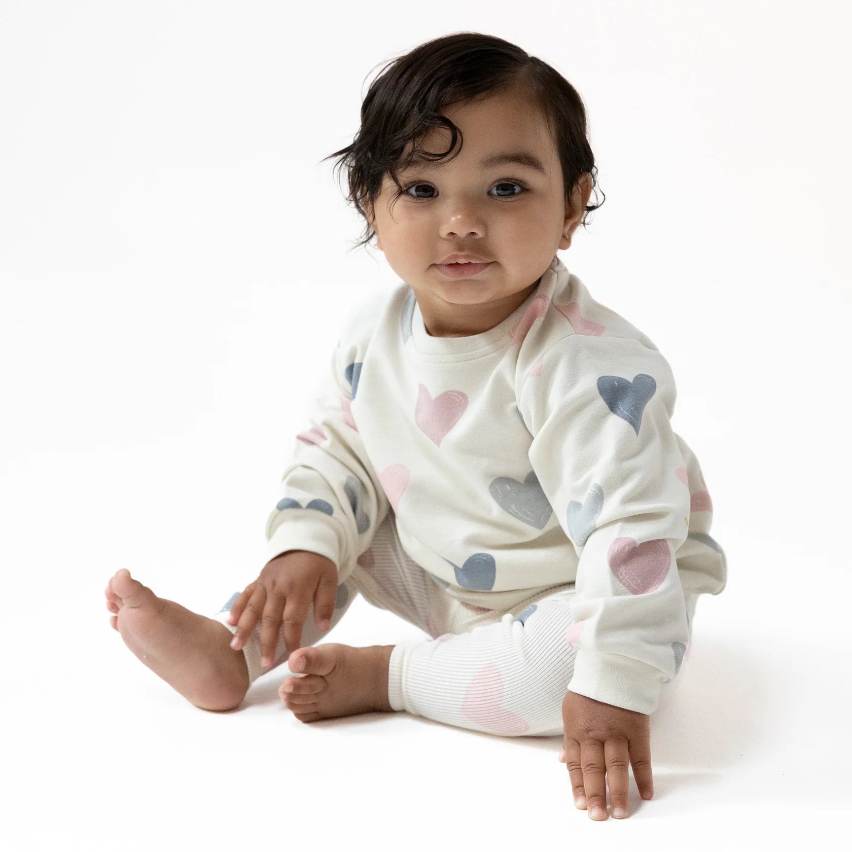 A baby sits on a white surface wearing the Angel Dear French Terry Puffy Long Sleeved Oversized Sweatshirt + Legging set with pink, blue, and gray heart patterns, looking toward the camera.