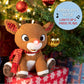 The Kids Preferred Rudolph Plush with Music and Lights, featuring a glowing nose and music, sits in front of a decorated holiday tree and gifts.