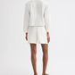 A person faces away, wearing a long-sleeve white jacket, TWP Cisco Short in coated viscose linen with pleats, and red sandals—a crisp summer look set against a plain white background.