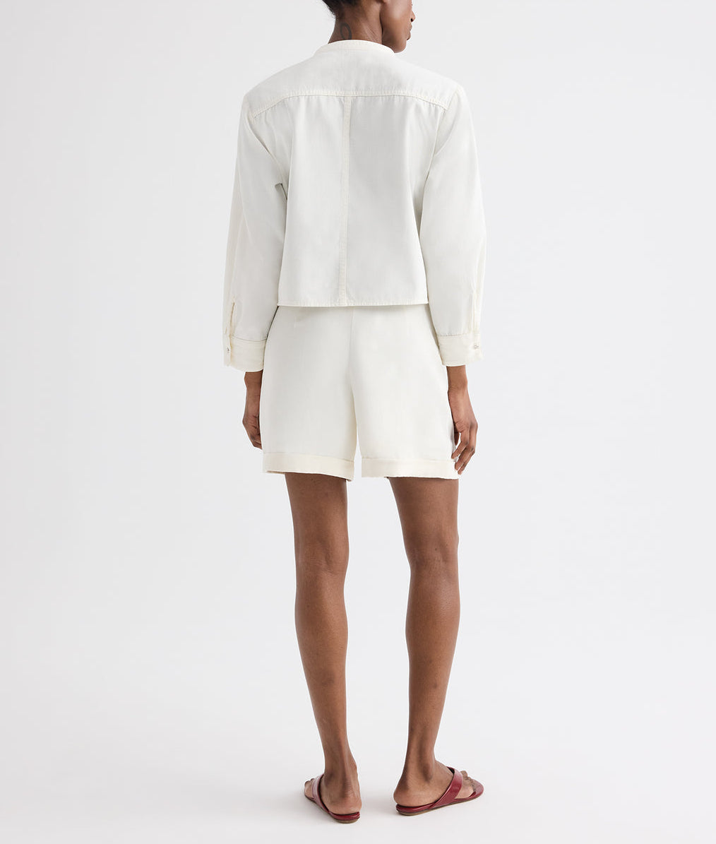 A person faces away, wearing a long-sleeve white jacket, TWP Cisco Short in coated viscose linen with pleats, and red sandals—a crisp summer look set against a plain white background.